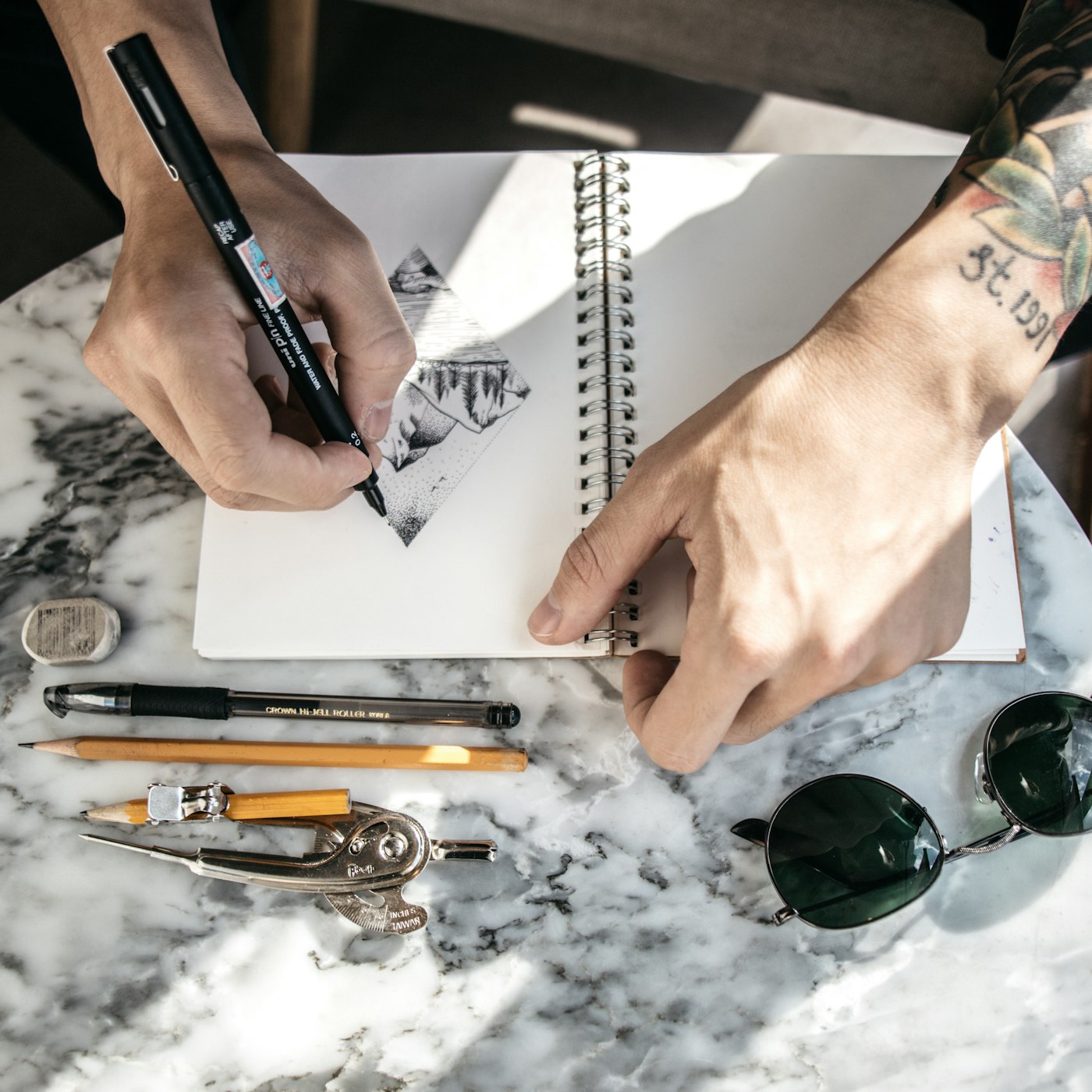 A hand sketching a design concept in a notebook, surrounded by pens, pencils, and drafting tools
